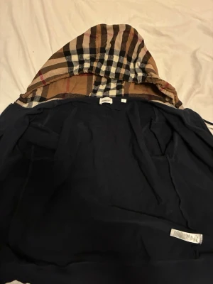 Marinblå hoodie med Burberry-huva - Snygg marinblå hoodie med dragkedja från Burberry. Huvan har det klassiska Burberry-rutiga mönstret i beige, svart, vitt och rött. Hoodien har fickor framtill och justerbar dragsko i huvan. Perfekt för dig som vill ha en stilren men ändå ikonisk look.