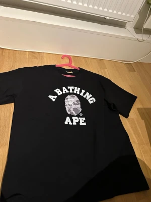 Svart BAPE t-shirt  - Säljer en svart t-shirt från A Bathing Ape med det klassiska BAPE-huvudet i grått camotryck och stor vit logga på bröstet. Använd 2-3 gånger men inga täcken på användning. Pris går att diskuteras. Öppen för frågor och bud. Mått kan skickas