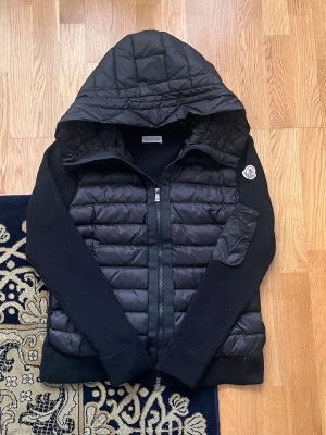 Svart pufferjacka från Moncler - Snygg svart pufferjacka från Moncler med quiltad front och rygg, stickade ärmar och huva. Jackan har dragkedja framtill, två sidofickor och en patch med Moncler-logga på ärmen. Perfekt för kalla dagar och riktigt trendig look.