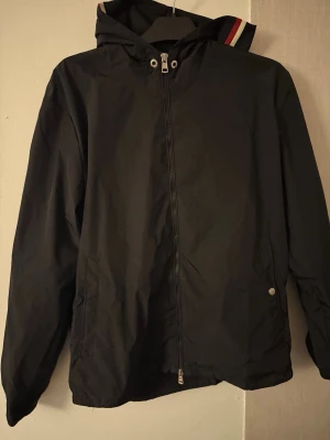 Svart vindjacka från Moncler - Snygg svart vindjacka från Moncler med huva och dragkedja framtill. Jackan har Moncler-logga på ärmen och detaljer i rött, vitt och blått på huvan. Ena knappen på fickan har lossnat, annars fel fri. Priset är inte hugget i sten bara att höra av sig. STORLEK 4 som motsvarar L