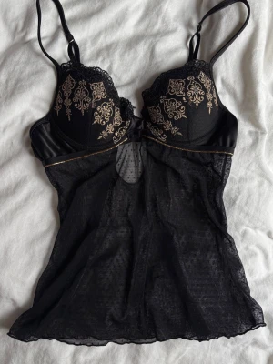 Svart topp med guldbroderi - Snygg svart topp med tunna axelband och vadderade kupor. Överdelen har vackra guldfärgade broderier och spetsdetaljer, medan nederdelen är i transparent mesh. Perfekt för dig som vill ha något elegant och unikt i garderoben.