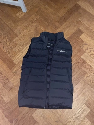 Svart dunväst från Sail Racing - Snygg svart dunväst från Sail Racing med hög krage och dragkedja framtill. Västen har två sidofickor och diskret logga på bröstet. Tillverkad i lätt och värmande material med 800 fill power dun. Perfekt för lager på lager under kyliga dagar.