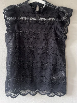 Svart spetsblus med volangärmar - Snygg svart blus i spets med volangdetaljer på axlarna. Blusen är ärmlös och har ett vackert spetsmönster över hela plagget samt vågig detalj nedtill. Aldrig använd endast prövad.