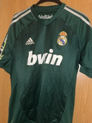 Grön Real Madrid tröja Adidas - Säljer en grön Real Madrid matchtröja från Adidas med vita detaljer och klubbmärke på bröstet. Tröjan har Bwin som sponsortryck och är tillverkad i lätt och ventilerande ClimaCool-material. Perfekt för dig som älskar fotboll och vill ha en unik supportertröja.