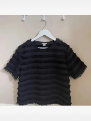 Svart randig topp med fransar - Cool svart topp med horisontella ränder och fransiga detaljer. Kortärmad och croppad modell med rund halsringning. Perfekt för dig som vill ha en unik och trendig look.