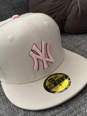 Beige New Era Yankees keps - Säljer en beige New Era 59FIFTY keps med rosa NY Yankees-logga framtill och broderade detaljer med blommor och motiv på sidan. Kepsen har rak skärm och klassisk snapback-look. Perfekt för dig som vill ha en stilren men ändå unik accessoar.