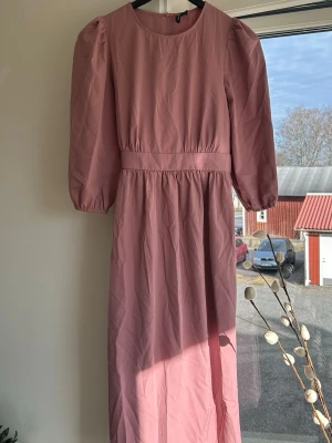 Lång rosa klänning med puffärm - Lång rosa klänning med puffärmar och rund halsringning. Klänningen har ett brett midjeband och ett knytband i ryggen för extra detalj. 💗