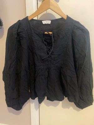 Svart blus med knytning från Mango - Svart blus från Mango med långärmad design och knytning framtill vid halsen. Blusen har en lös passform, lätt puffade ärmar och fina spetsdetaljer upptill. Tillverkad i ett tunt, mjukt tyg som ger en avslappnad look.