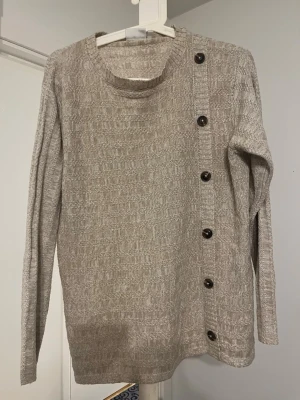 Beige stickad tröja med knappar - Stickad beige tröja med rund hals och långa ärmar. Unik detalj med svarta knappar längs ena axeln och sidan framtill. Tröjan har ett diskret mönster i stickningen och är i mjukt material, perfekt för lager på lager.
