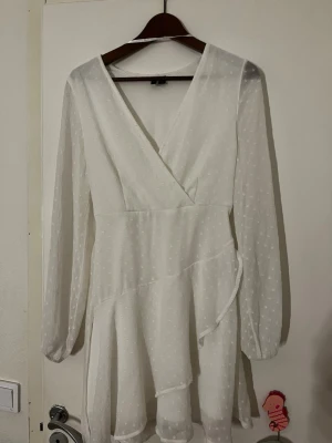 Vit omlottklänning med prickar - Söt vit kortklänning med omlottmodell och v-ringning. Klänningen har långa, transparenta ärmar och ett diskret prickigt mönster i tyget. Volangdetaljer nedtill och knyt i midjan ger en feminin look. Perfekt för sommarens alla tillfällen.