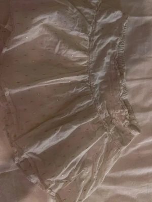 Vit glittrig kortkjol med resår - Söt vit kortkjol med glittriga detaljer och små prickar. Kjolen har resår i midjan för en bekväm passform och är gjord i ett lätt material som ger en luftig känsla. Perfekt för dig som vill ha en feminin och trendig look.