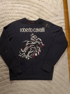 Roberto Cavalli sweatshirt  - Lila sweatshirt från Roberto Cavalli med stort vitt logotryck och en grafisk tiger på bröstet vitt, svart och vinrött. Tröjan har rund halsringning, ribbade muddar och är tillverkad i mjuk materialsblandning av cotton och polyester. Perfekt för en trendig och avslappnad stil. Tags och kvitto kommer med. 