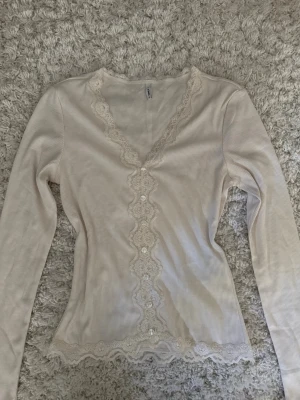 Spetsblus - Söt beige långärmad blus med v-ringning, spetsdetaljer längs knäppningen och nederkanten samt små knappar framtill. Tunn ribbad struktur och figurnära passform. Perfekt för dig som gillar romantiska detaljer.