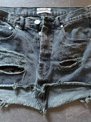Svarta slitna jeansshorts One Teaspoon W28 - Riktigt snygga svarta jeansshorts från One Teaspoon med slitna detaljer och råa kanter. Shortsen har hög midja, klassisk femficksdesign och stora slitningar framtill för en edgy look. Perfekta till sommaren för en avslappnad stil.