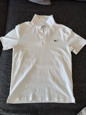 Vit pikétröja från Lacoste - Klassisk vit pikétröja från Lacoste med korta ärmar och den ikoniska gröna krokodilloggan broderad på bröstet. Tröjan har krage och knappar vid halsen. Perfekt till en clean och stilren look.