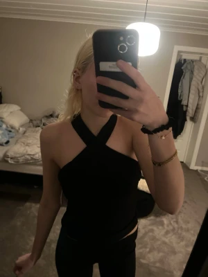 Svart omlott halterneck topp - Snygg svart topp med omlott halterneck-design och ärmlös passform. Toppen har en tight siluett och korsade axelband framtill som ger en trendig look. Perfekt att styla med jeans eller kjol för en cool outfit.