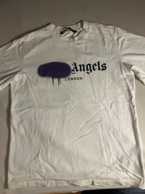 Vit Palm Angels t-shirt med tryck - Säljer en vit t-shirt från Palm Angels med ikoniskt tryck framtill. T-shirten har svart text och en lila spray paint-effekt över loggan. Klassisk passform och rund hals. Perfekt för dig som gillar streetwear och vill sticka ut. Nästan som ny knappt använt 