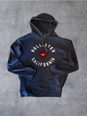 Hollister hoodie  - Mörkblå hoodie från Hollister med stor huva och dragsko. På bröstet finns ett broderat rött och vitt Hollister California-märke och klassisk fågel-logga. Hoodien har känguruficka framtill och mjukt material på insidan för extra komfort. Bra i passform!