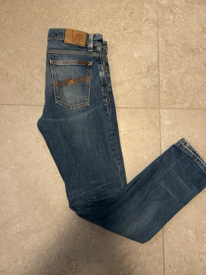 Blå jeans från Nudie Jeans - Säljer ett par klassiska blå jeans från Nudie Jeans med snygga kontrastsömmar och den ikoniska vågdetaljen på bakfickan. Jeansen har normal passform och är gjorda i slitstark denim.