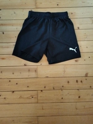 Svarta shorts från Puma - Svarta shorts från Puma med elastisk midja och klassisk logga i vitt på vänster ben. Perfekta för träning eller chill, tillverkade i ett lätt och bekvämt material som andas. Enkel och stilren design som funkar till det mesta. Orginal pris 250 kr
