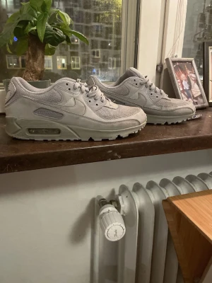 Nike Air Max 90 grå sneakers - Säljer ett par stilrena Nike Air Max 90 sneakers i helgrå färg. Skorna har en klassisk siluett med mesh och mockadetaljer, samt den ikoniska synliga Air-dämpningen i sulan. Perfekta för dig som gillar en clean och sportig look.