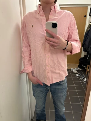 Rosa skjorta från Ralph Lauren - Klassisk rosa skjorta från Ralph Lauren med button-down krage och broderad logga på bröstet. Skjortan har långa ärmar, knappar framtill och är tillverkad i mjuk bomull. Perfekt för en clean och stilren look.
