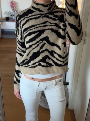 Beige och svart zebramönstrad stickad tröja - Säljer en stickad tröja med zebramönster i beige och svart. Tröjan har hög hals, långa ärmar och ribbade muddar vid ärmslut och nederkant