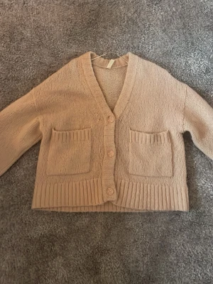 Beige stickad kofta med fickor - Mysig beige stickad kofta med v-ringning och tre knappar framtill. Koftan har två stora fickor på framsidan och ribbade muddar vid ärmslut och nederkant. Perfekt för lager på lager och enkel att matcha.