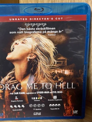 Drag Me To Hell Blu-ray  - Blu-ray-utgåva av skräckfilmen Drag Me To Hell i Unrated Director's Cut. Omslaget är svart och rött med motiv av en kvinna och eld. Filmen är regisserad av Sam Raimi och har flera svenska recensioner på framsidan. Perfekt för dig som gillar skräck och spänning.