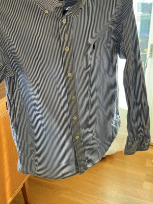 Blåvit randig skjorta Ralph Lauren - Snygg blåvit randig skjorta från Ralph Lauren med klassisk krage, knappar framtill och lång ärm. Skjortan har en liten broderad logga på bröstet och är gjord i mjuk bomull för en bekväm känsla. Perfekt för en clean och stilren outfit.