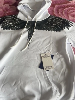 Vit hoodie Marcelo Burlon med vingar - Snygg vit hoodie från Marcelo Burlon County of Milan med ikoniskt svart vingtryck över axlar och bröst. Hoodien har huva med dragsko och stor magficka framtill. Tillverkad i mjukt material som känns skönt mot huden. Perfekt för dig som vill sticka ut med en unik design.