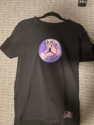 Svart PSG x Jordan t-shirt - Snygg svart t-shirt från Jordan i samarbete med Paris Saint-Germain. Stor logga på bröstet i lila och rosa toner med Jordan-loggan i mitten. Klassisk rund hals och korta ärmar. Perfekt för dig som gillar streetwear och sportig stil.