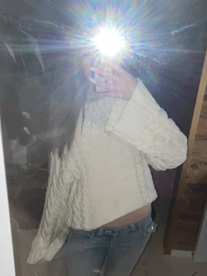 Vit kabelstickad tröja - Säljer en vit kabelstickad tröja med breda flätmönster längs ärmarna och sidorna. Tröjan har en avslappnad passform, ribbad rund halsringning och långa ärmar. Perfekt för lager på lager och riktigt mysig till jeans.