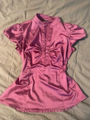 Rosa satinblus från Savida - Snygg rosa blus från Savida i glansigt satinmaterial. Blusen har korta puffärmar, ståkrage och dekorativa knappar framtill med rynkade detaljer vid bysten. Perfekt för dig som vill ha en feminin och trendig look. Står strl 36 men är en 38 