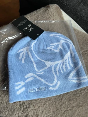Ljusblå mössa från Arc'teryx - Snygg ljusblå mössa från Arc'teryx med vit logga och stort grafiskt mönster på framsidan. Tillverkad i mjukt, stretchigt material som sitter skönt på huvudet. Perfekt för dig som vill ha en stilren och sportig look.