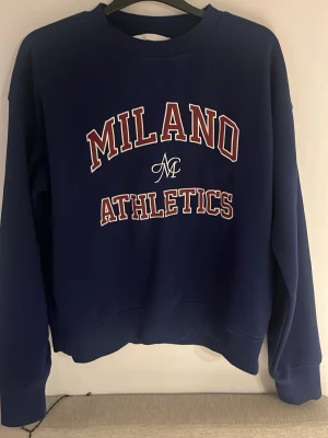 Blå sweatshirt  H&M - Mörkblå sweatshirt från H&M med trycket 'Milano Athletics' i vitt och rött på bröstet. Tröjan har rund halsringning, långa ärmar och ribbade muddar vid ärmslut och nederkant. Perfekt för en sportig och avslappnad stil. Använd 1 gång 