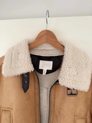 Pilotjacka från H&M - Jättefin och mysig pilotjacka från H&M i storlek M. Jackan är gjord i mjuk mockaimitation med ett varmt och fluffigt foder i teddy/sherpa. Den är perfekt för höst och mildare vinterdagar.  Använd under en säsong men i mycket gott skick utan tydliga tecken på slitage. En klassisk modell som aldrig går ur tiden och passar till både jeans och klänning. Fins att hämta i Kungsängen eller Sundbyberg. 
