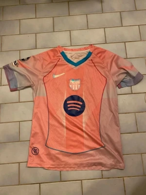 Rosa FC Barcelona matchtröja Nike - Säljer en rosa och ljuslila FC Barcelona matchtröja från Nike med klubbmärke och Spotify-logga på bröstet. Tröjan har korta ärmar, V-ringning med turkos kant och är tillverkad i ett lätt, ventilerande material perfekt för träning eller match.
