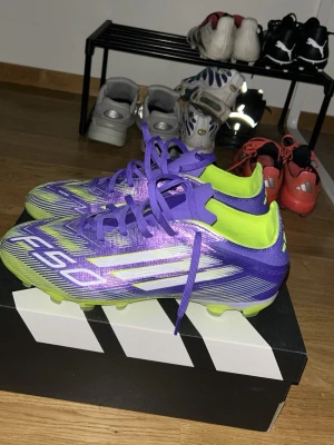 Adidas F50 lila fotbollsskor - Säljer ett par Adidas F50 fotbollsskor i en riktigt snygg lila och neongul färgkombination. Skorna har klassiska vita stripes på sidan, snörning och FG-dobbar för spel på naturgräs. Tillverkade i syntetmaterial för lätt vikt och bra bollkontroll. Perfekta för dig som vill sticka ut på planen!