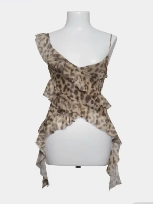 Leopardmönstrad volang topp - Säljer en leopardmönstrad linnetopp med volangdetaljer och asymmetrisk design. Toppen har tunna axelband och är tillverkad i ett lätt, transparent material som ger en luftig känsla. Perfekt för dig som vill sticka ut med en edgy och trendig look.