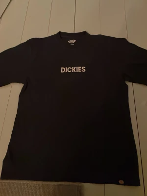 Svart Dickies t-shirt med tryck - Snygg svart t-shirt från Dickies med stort vitt logotryck framtill och en cool grafisk print i orange och vitt på ryggen. Klassisk passform och rund hals. Perfekt för dig som gillar streetwear och vill ha något stilrent men ändå med attityd.