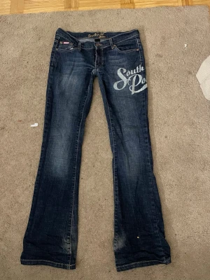 Low waist jeans - Dom har tyver blivigt små men dom är mer bootcut när dom passar bättre!🥰Säljer ett par mörkblå bootcut jeans från Southpole med stor vit logga på ena låret. Jeansen har låg midja, klassiska fem fickor och slitningar för en cool vintagekänsla. Skitsnygga och y2k/alternativ stil. Jag har sytt ner dom lite så dom blir mer low waist! Skriv för frågor och annat!💕💕🥰