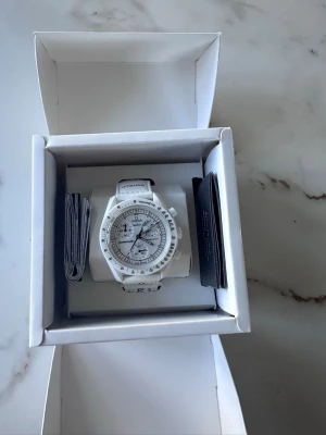 Vit Omega x Swatch Speedmaster - Säljer en stilren vit Omega x Swatch Speedmaster MoonSwatch med tachymeter och kronograf. Klockan har vit boett och matchande vitt band med svart text. Snygg och modern design som sticker ut på handleden.