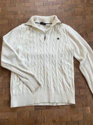 Vit kabelstickad tröja från Morris - Kabelstickad vit tröja från Morris med halv dragkedja och ribbad krage. Tröjan har ett diskret Morris-emblem broderat på bröstet och ribbade muddar vid ärmslut och nederkant. Perfekt för lager på lager och klassisk stil. Tröjan har en väldigt liten prick på vänster bakre axel(se bild 4), men inget som man märker av. Superbra skick, använd ett fåtal gånger. Nypris ca 2000