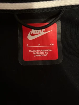 Nike Tech Fleece hoodie svart/grå - Nike Tech Fleece hoodie i svart och grått med dragkedja framtill och en grön Nike-logga på bröstet. Hoodien har en huva, ficka på ärmen med dragkedja och sportig design. Perfekt för dig som gillar streetwear och vill ha en stilren look.