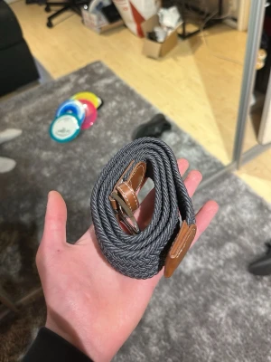 Grått flätat bälte med läderdetaljer - Snyggt grått flätat bälte med bruna läderdetaljer och silverfärgat spänne. Bältet har en stilren design och passar perfekt till jeans eller chinos. Flexibelt material som gör det enkelt att justera längden efter behov.   