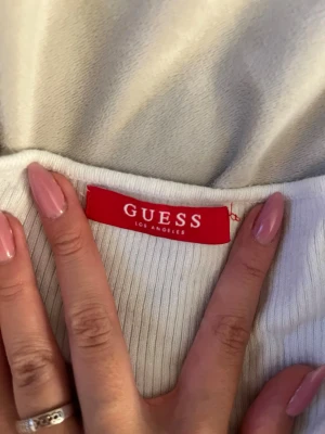 Vit ribbad långärmad topp Guess - Snygg vit ribbad långärmad topp från Guess. Modellen är figurnära med stretchigt material och har en enkel, stilren design som passar till det mesta. Perfekt för dig som gillar klassiska och trendiga plagg från populära märken.