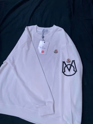 Vit sweatshirt från Moncler - Vit sweatshirt från Moncler med rund halsringning, ribbade muddar och stort svart Moncler-logotryck på bröstet. Tröjan har även en broderad patch ovanför loggan. Tillverkad i mjukt material för en clean och modern look. Det står att det är M men den passar mig som är S
