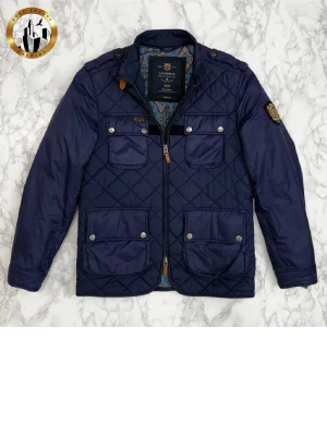 Mörkblå quiltad jacka Morris - Snygg mörkblå quiltad jacka från Morris med fyra framfickor med tryckknappar, axelklaffar och patch på ärmen. Jackan har dragkedja framtill och detaljer i brunt läder vid kragen. Fodret har ett färgglatt mönster som ger extra stilpoäng.