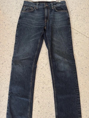 Mörkblå jeans från Nudie Jeans - Säljer ett par mörkblå jeans från Nudie Jeans med klassisk rak passform och snygga slitningar. Jeansen har orangea sömmar och är tillverkade i slitstark denim. Perfekta för dig som gillar en tidlös och stilren look. Helt oanvända.